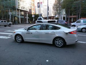 A white Uber car at an intersection raised the question of who pays when a rideshare driver causes auto accident damage to your vehicle.