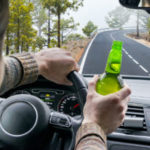 Drunk Young Man Driving A Car On The Road With A Bottle Of Beer.